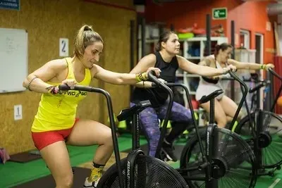 Un hombre levanta dos pesas rusas en un gimnasio con grandes ventanales y maquinaria industrial al fondo.