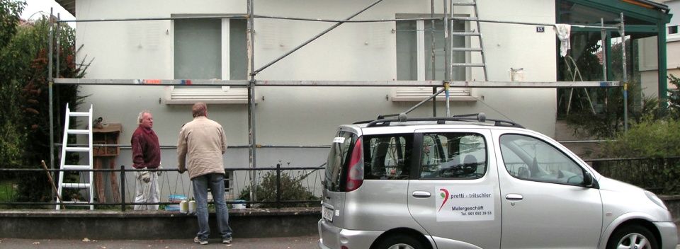 Zwei Männer an einem Gebäude mit Baugerüst, einer weiteren Leiter und einem davor geparkten Auto.