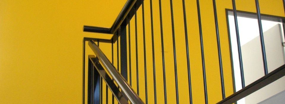 Gelbe Wand mit schwarzem Geländer und Metallhandlauf. Fenster sichtbar.