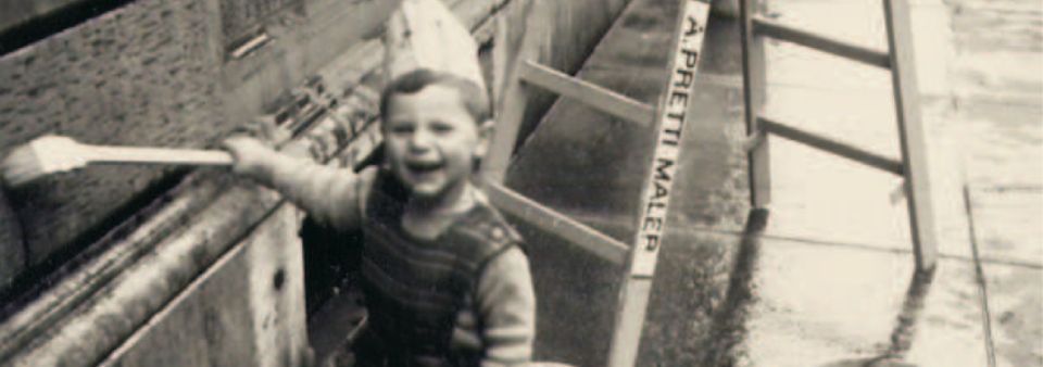 Ein kleines Kind mit einem breiten Grinsen hält eine Bürste. Es befindet sich im Freien neben einer Leiter.