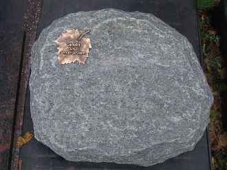 Dunkelgrauer, unregelmäßig geformter Grabstein mit Bronze-Blumenornament.
