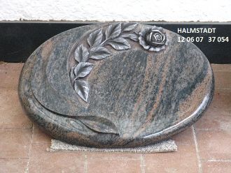 Ovaler Grabstein in Grün-Grau mit polierter Oberfläche und floralem Relief oben links.