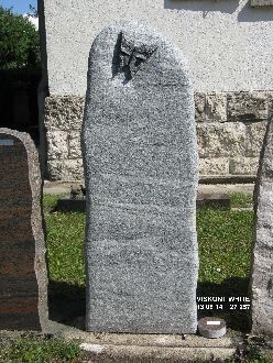 Grauer, hoher Grabstein mit einer eingravierten Taube oben links, steht auf einem Friedhof vor einer Steinmauer