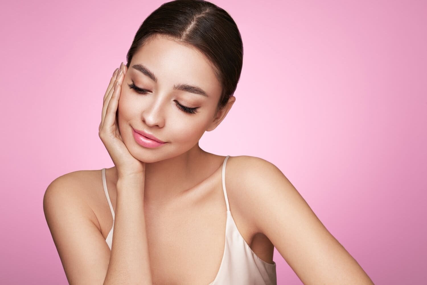 Una persona con la mano tocándose suavemente la mejilla sobre un fondo rosa liso, mirando hacia abajo con una suave sonrisa.