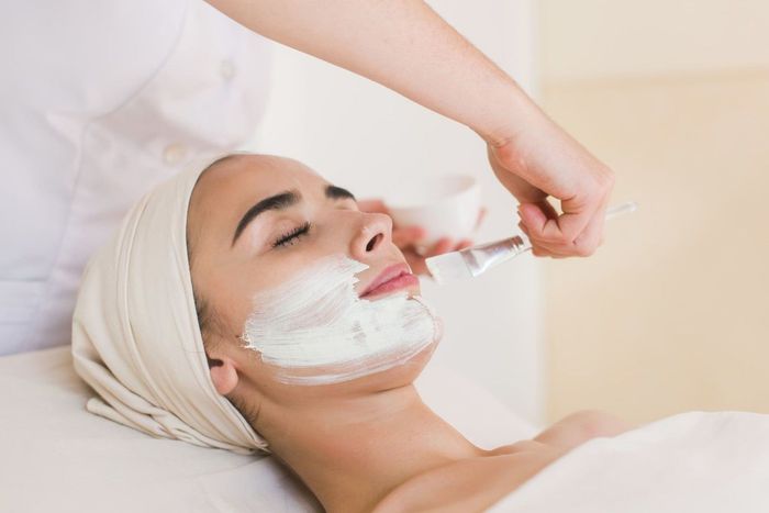 Una persona tumbada en una camilla de spa recibe la aplicación de una mascarilla facial por parte de un profesional.