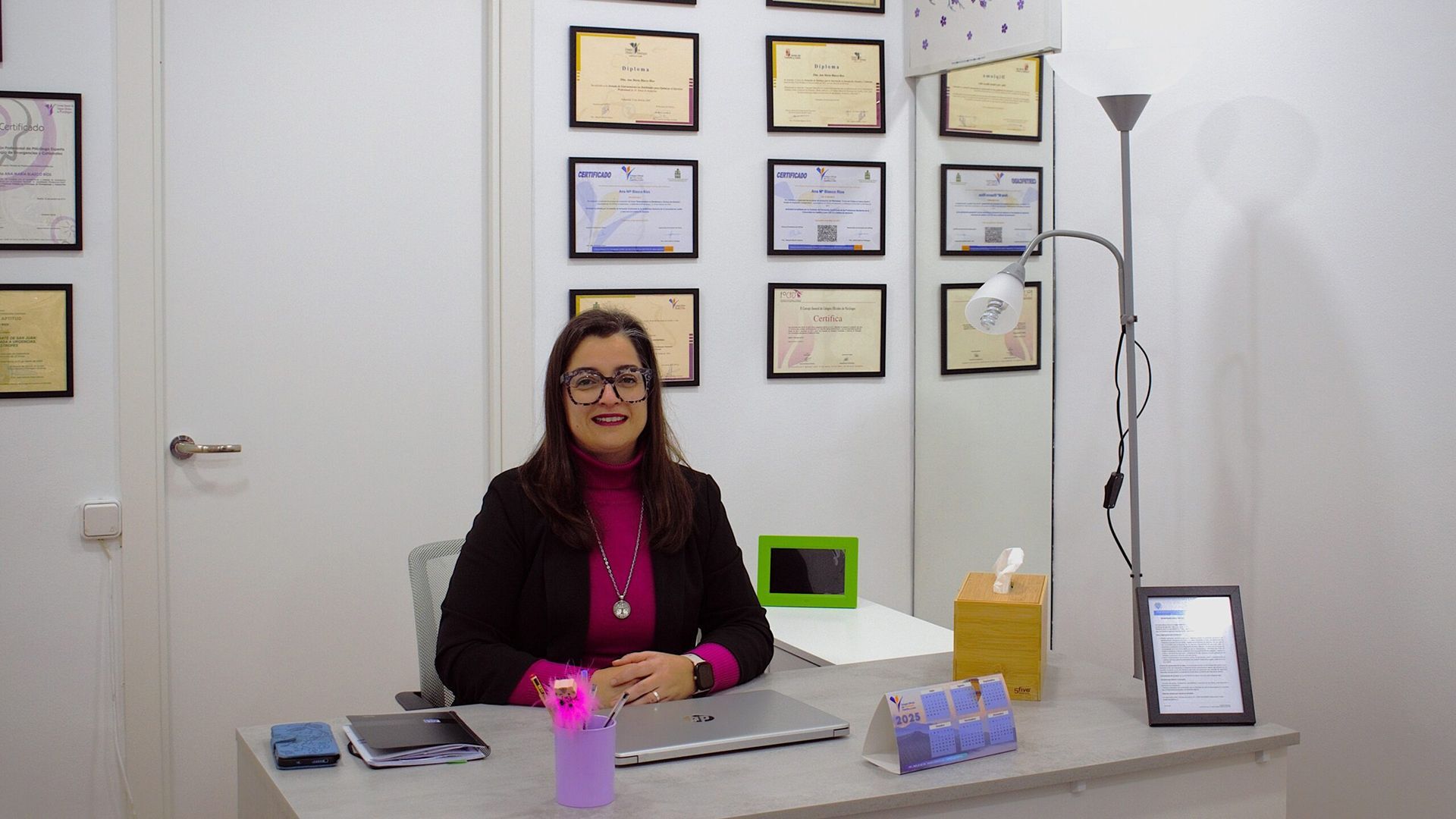 Mujer en la oficina, sonriendo, sentada en el escritorio con una computadora portátil, certificados enmarcados en la pared.