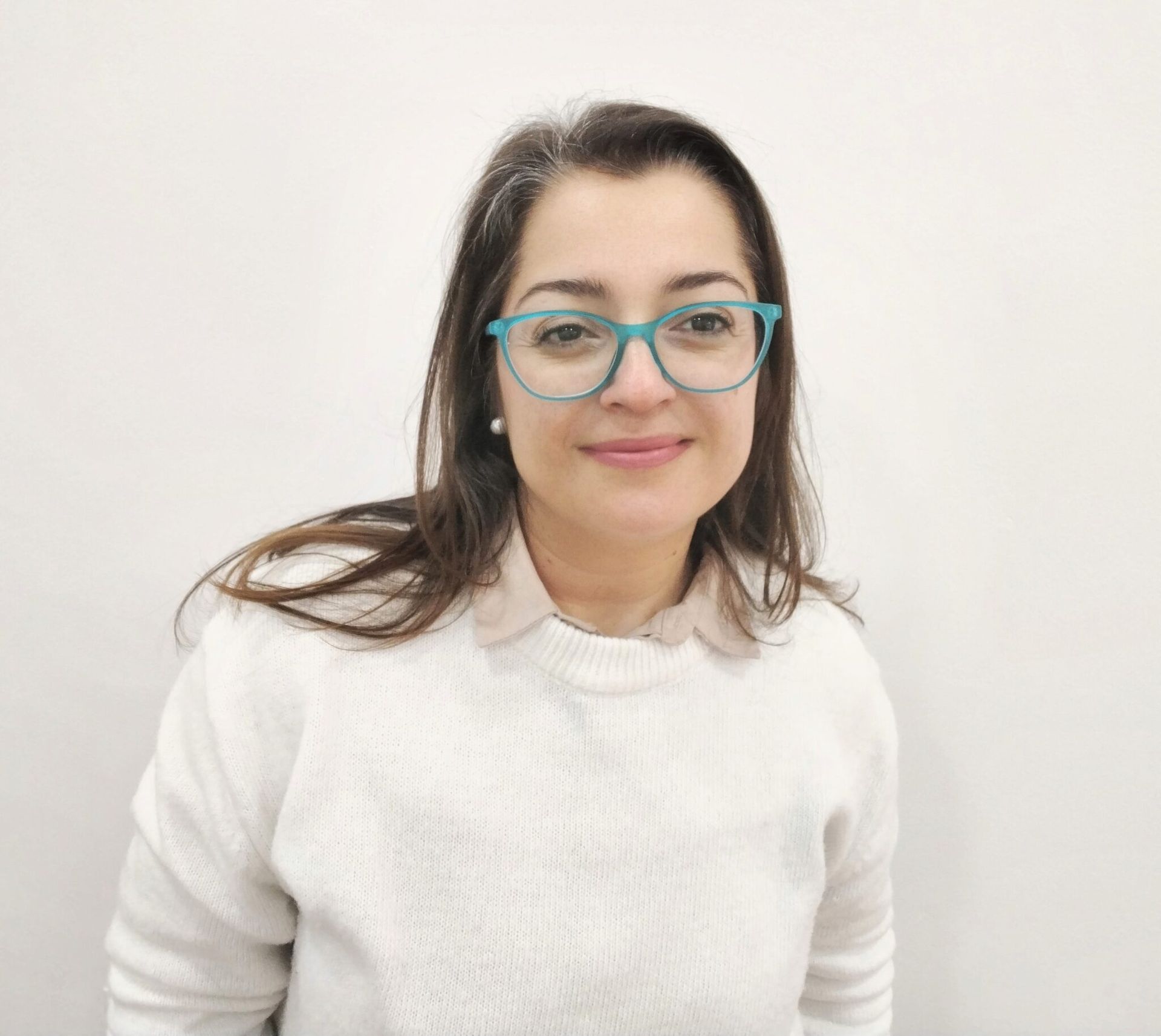 Una mujer con gafas y un suéter blanco sonríe a la cámara contra una pared blanca.