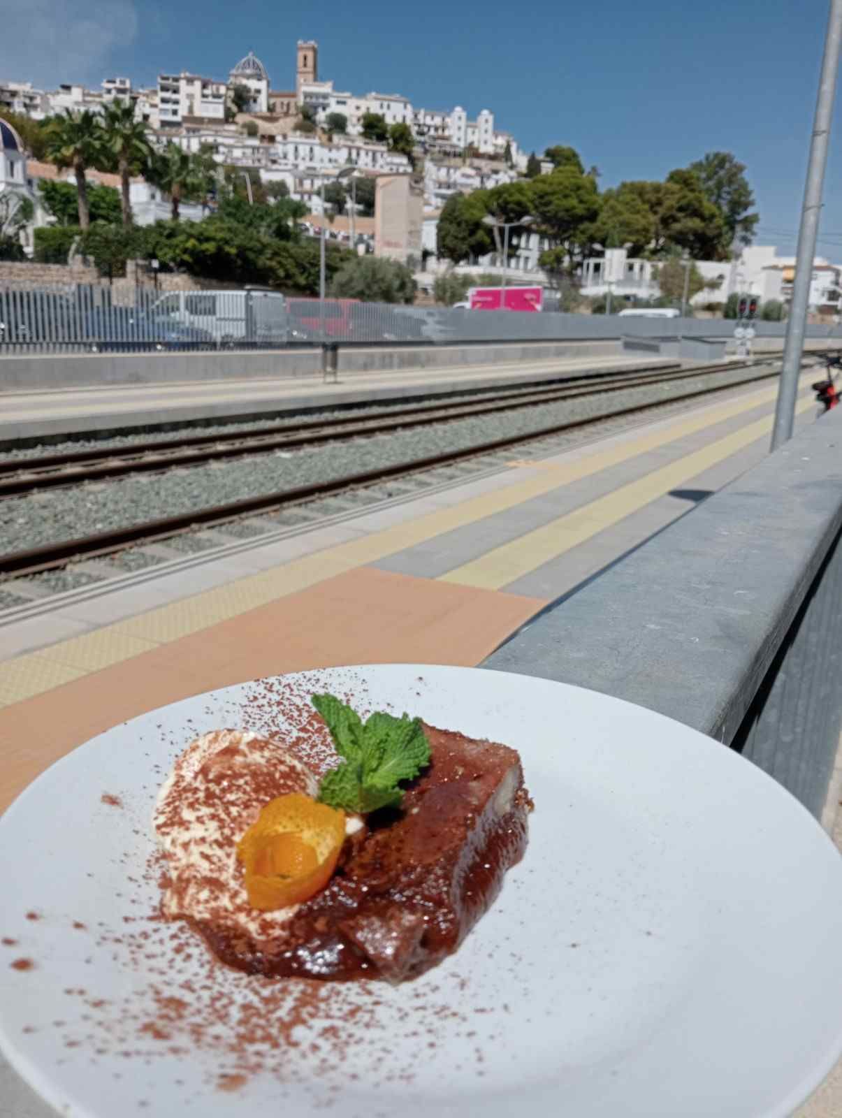 Postre en un plato blanco en el andén de una estación de tren, con las vías y un pueblo en la ladera al fondo.