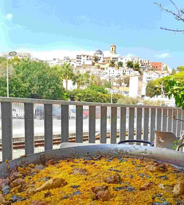 Pétalos de flores en una fuente circular, con un pueblo en la ladera y el cielo azul al otro lado de una barandilla.