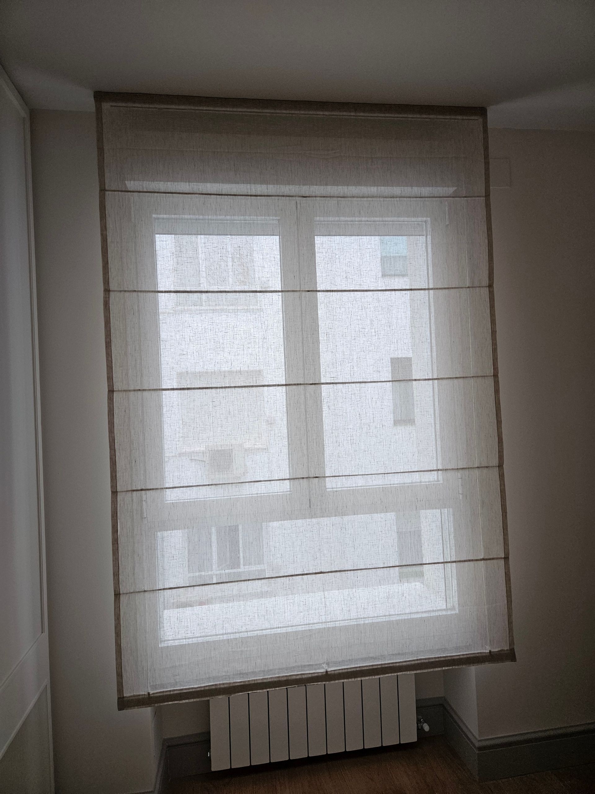 Cortina romana de color beige que cubre una ventana en una habitación con un radiador debajo.