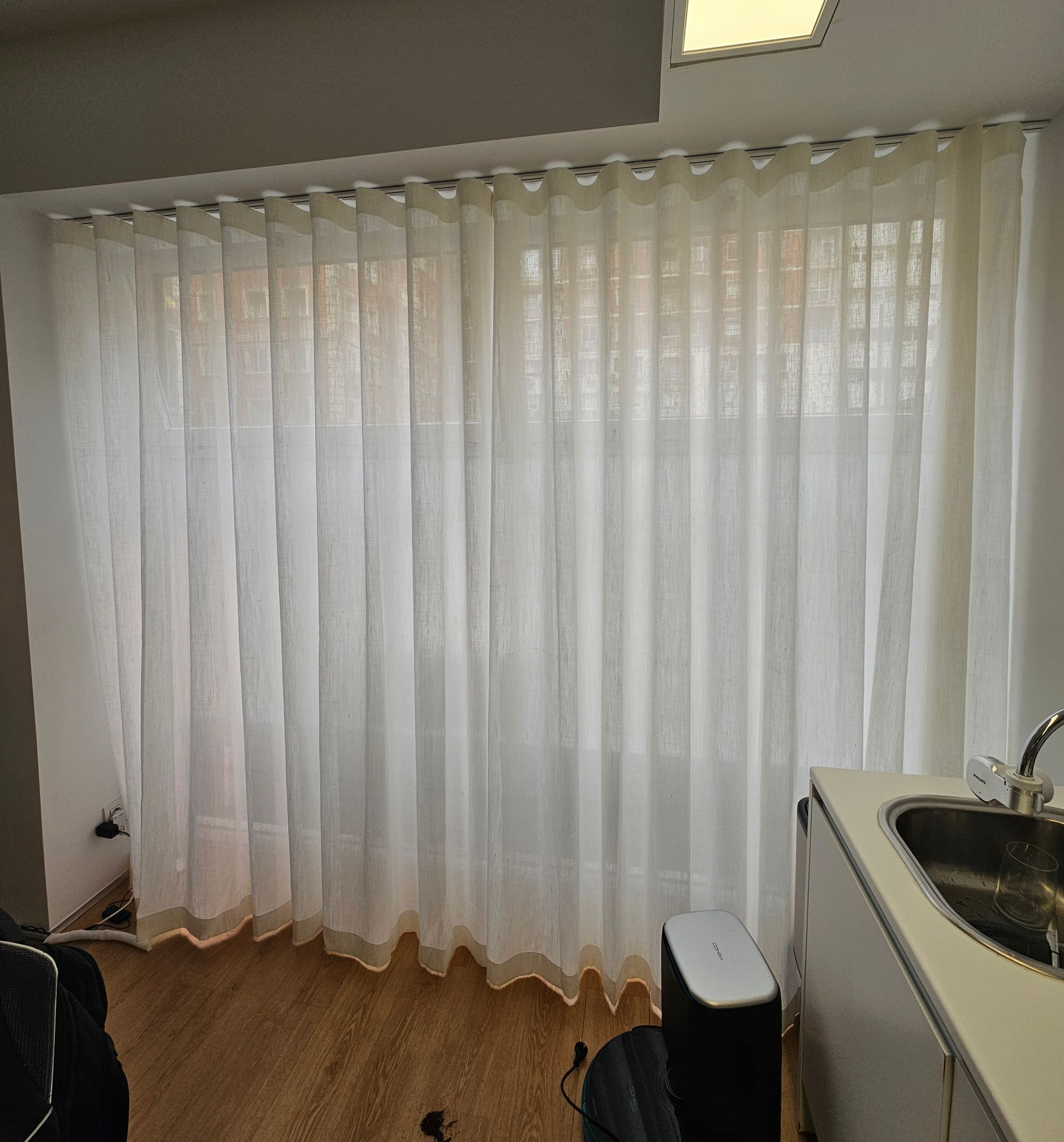Cortinas blancas cubren una ventana, cerca de un lavabo y una encimera, en una habitación con suelo de madera.