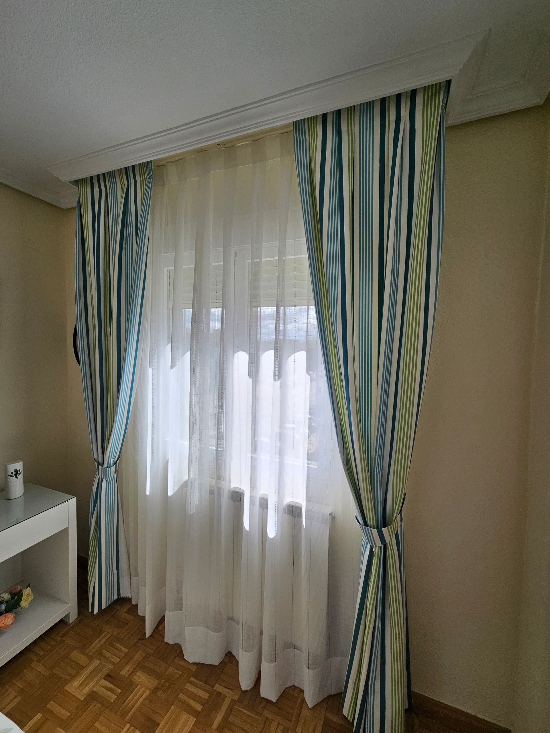 Ventana del dormitorio con cortinas blancas transparentes y cortinas a rayas azules y verdes recogidas.