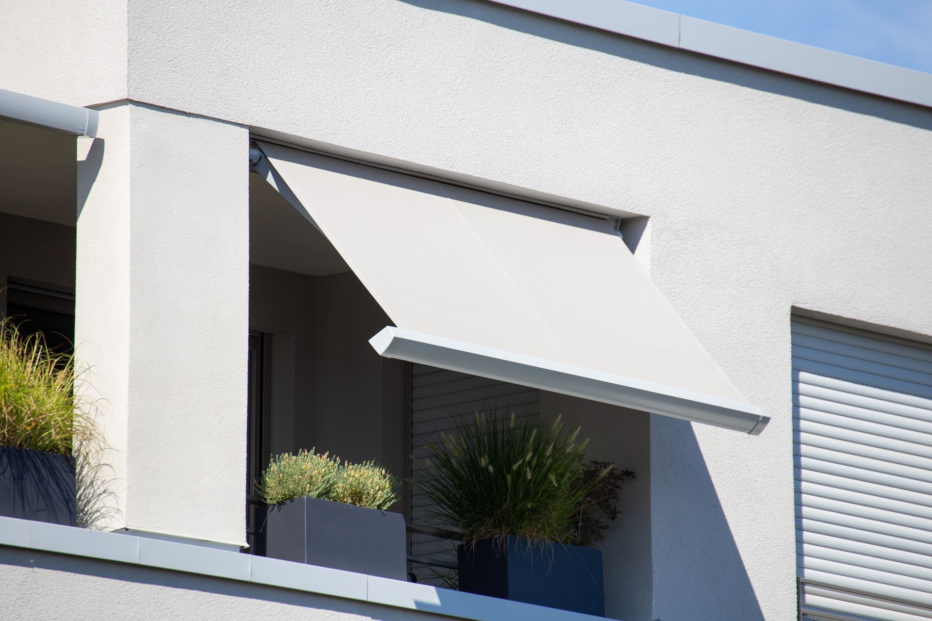 Balkon mit ausgefahrener grauer Markise, Pflanzen in quadratischen Pflanzkübeln und einem weißen Gebäude.
