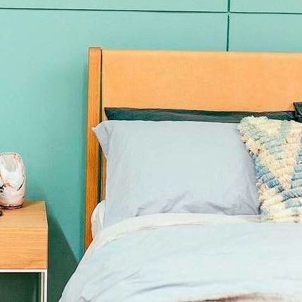 Dormitorio con ropa de cama azul claro, cabecero de madera y pared turquesa. Una mesita de noche sostiene un jarrón.