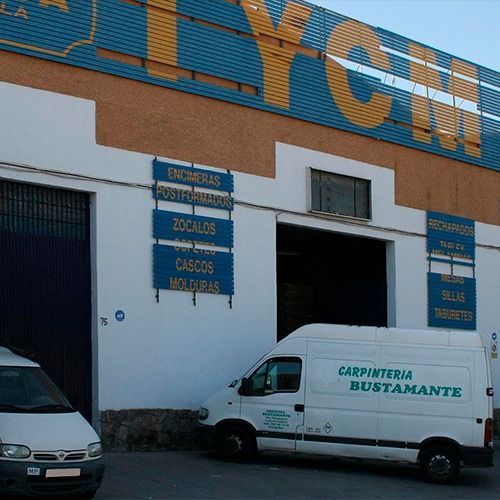 Exterior de un taller de carpintería con señalización azul y blanca; furgonetas blancas estacionadas afuera.