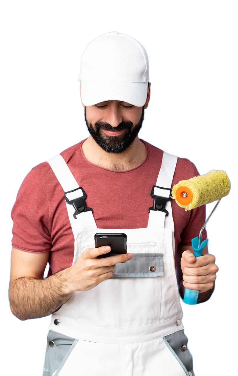 Un homme en salopette et casquette blanches qui tient un rouleau à peinture de la main gauche et regarde un téléphone de la main droite