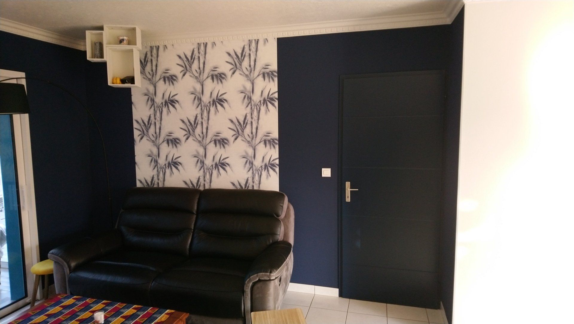 Pièce sombre aux murs bleu marine, papier peint à motifs de feuilles de palmier, canapé en cuir noir
