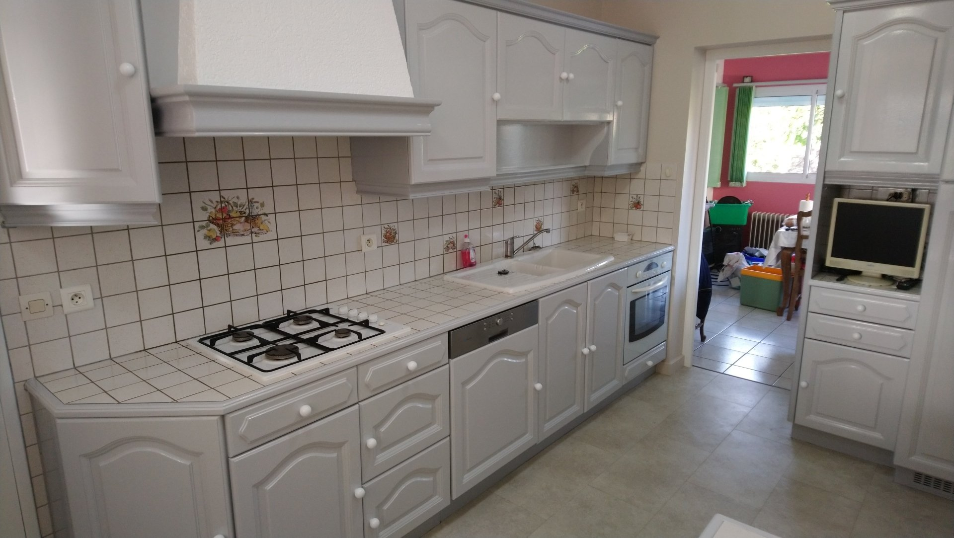 Cuisine blanche lumineuse avec une crédence carrelée, une cuisinière à gaz, un four encastré et une porte ouverte sur une pièce colorée