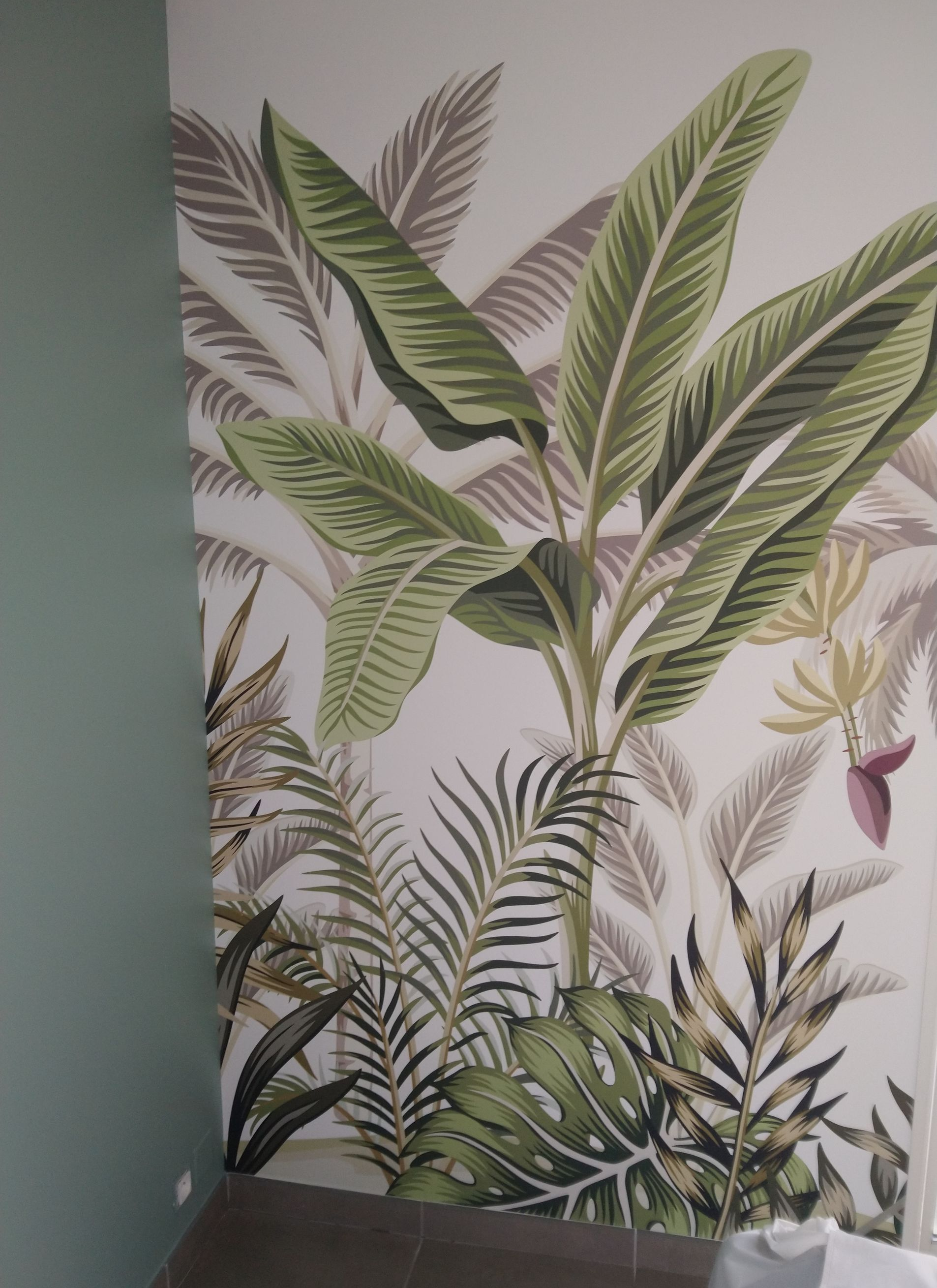 Papier peint mural à motif de feuilles tropicales dans des tons verts, crème et roses, sur un coin de mur