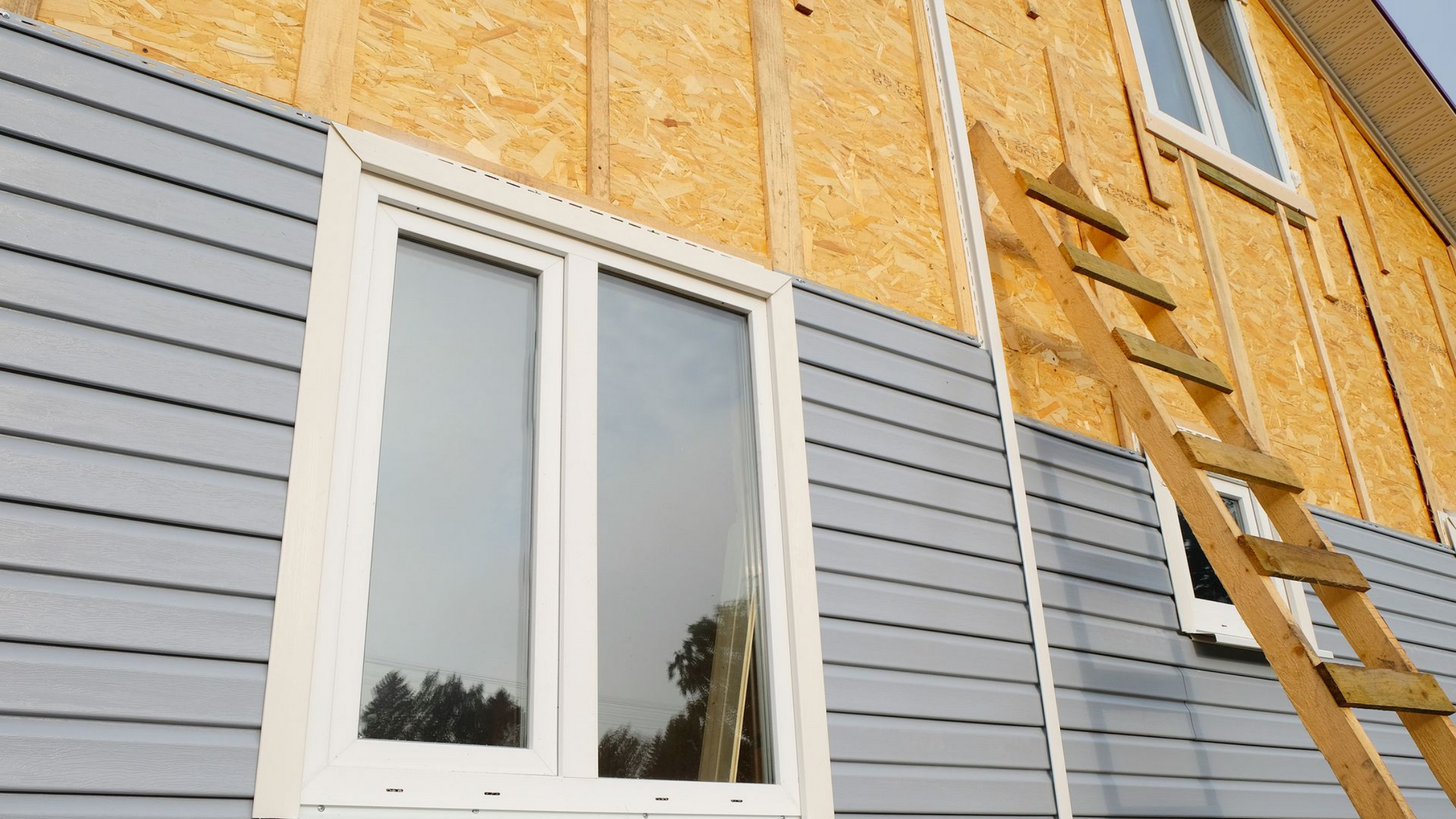Mur extérieur avec fenêtre blanche, bardage gris, revêtement en bois apparent et une échelle contre le mur.