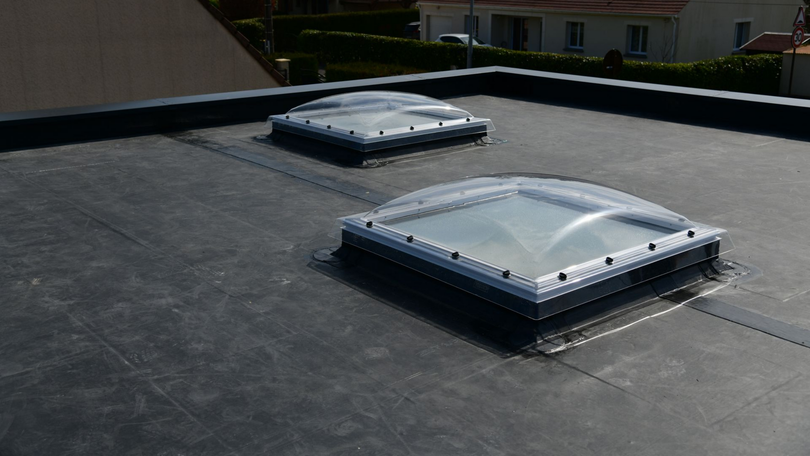 Deux puits de lumière sur un toit plat noir, avec des maisons visibles en arrière-plan.