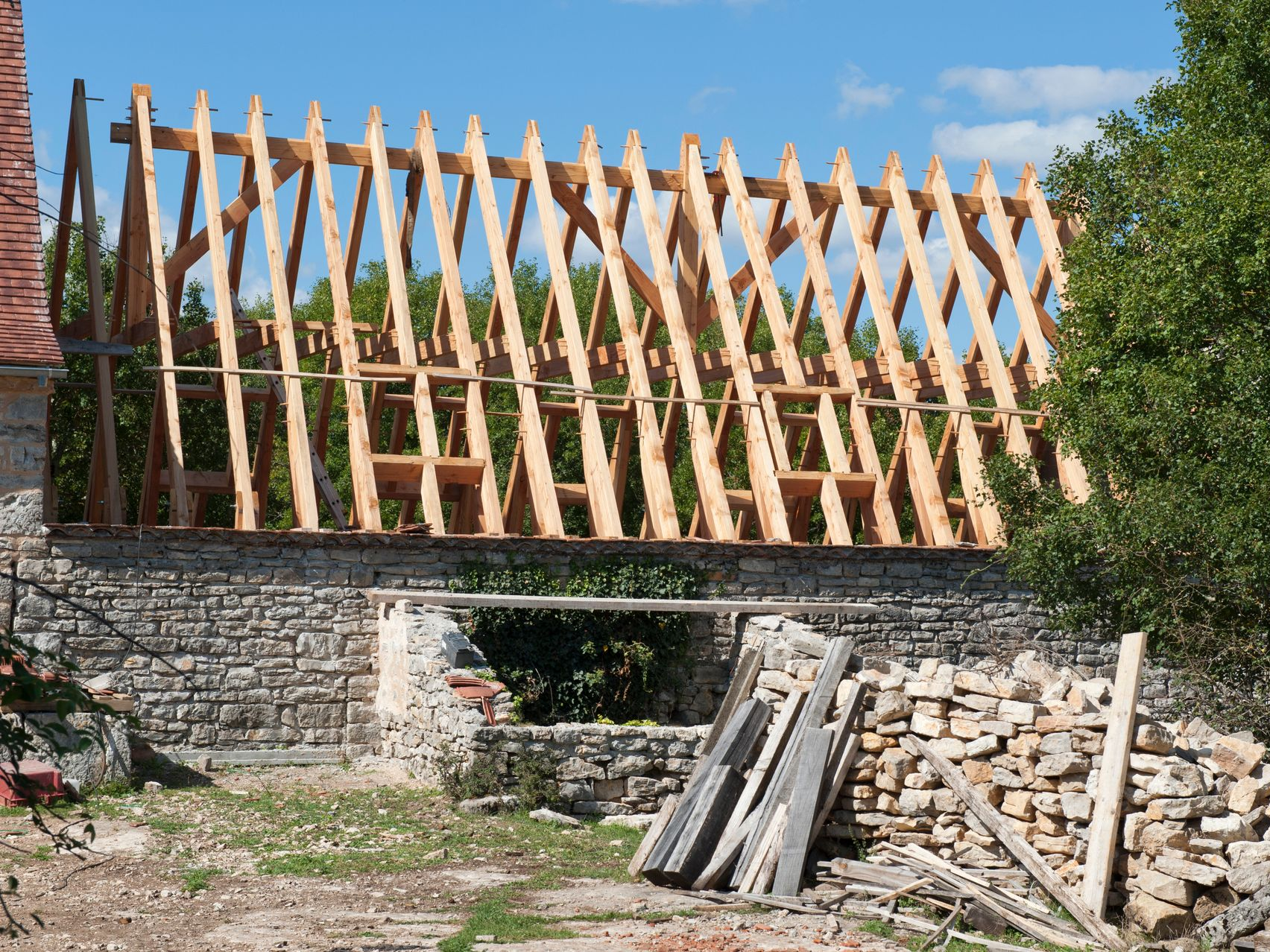 Charpente de toit en bois en construction au sommet d'un bâtiment en pierre, avec du bois empilé à proximité.