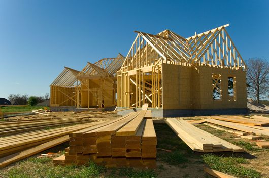 Maisons à ossature bois en construction sur un terrain ensoleillé, avec du bois empilé au premier plan.