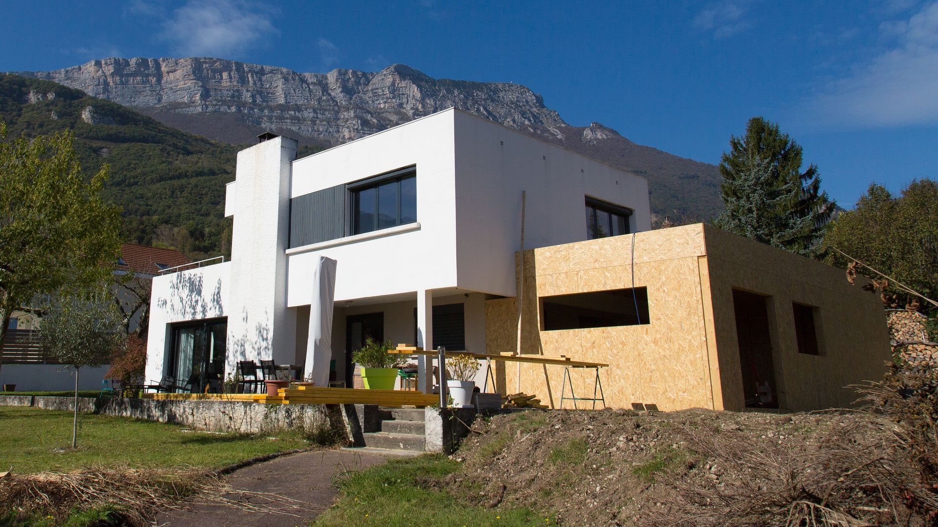 Maison blanche moderne avec extension en bois dans un cadre montagneux.