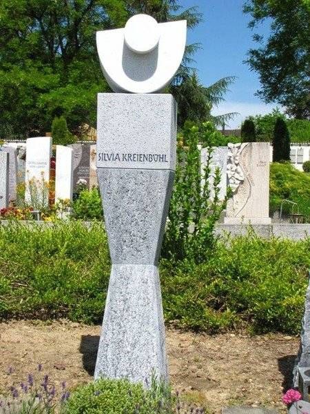 Weisses abstraktes Denkmal auf einem Steinsockel in einem grünen Friedhof oder Gedenkpark.