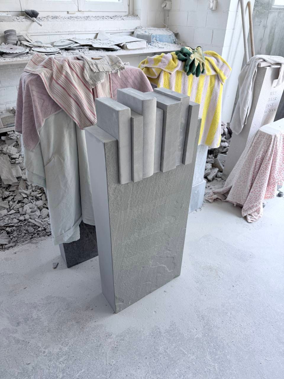 Graue Betonplatte mit vier vertikalen Lamellen in einer hellen Werkstatt, drapiert mit Tüchern und Handtüchern.