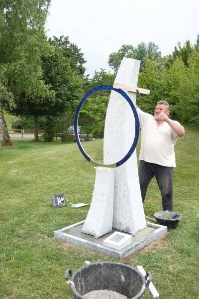Ein Mann bemalt im Freien auf einer Rasenfläche eine blaue Ringskulptur; Farbeimer stehen daneben.