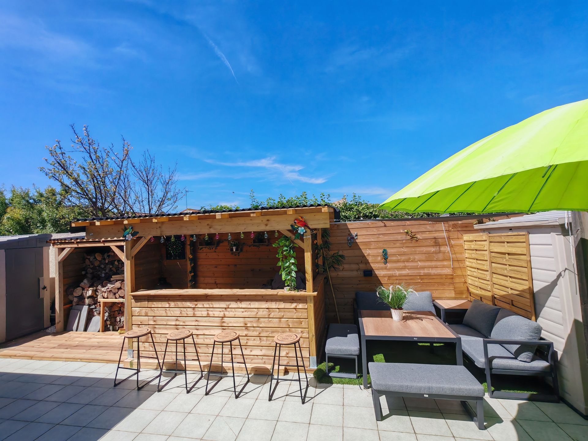 Terrasse arrière avec bar en bois, sièges gris et parasol vert vif sous un ciel bleu