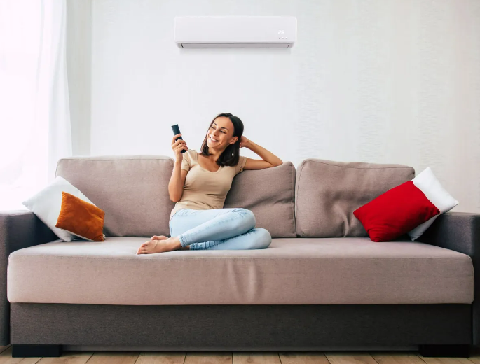 Une femme se détend sur un canapé gris, tenant une télécommande, avec des coussins rouges et orange dans un salon lumineux.