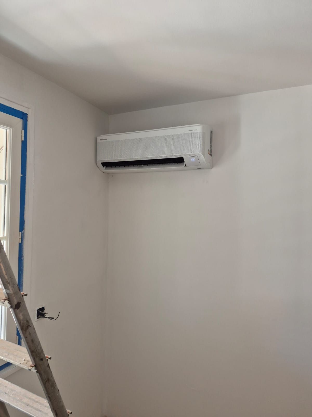 Climatiseur mural blanc avec échelle et bord de fenêtre recouvert de ruban adhésif bleu sur la gauche.