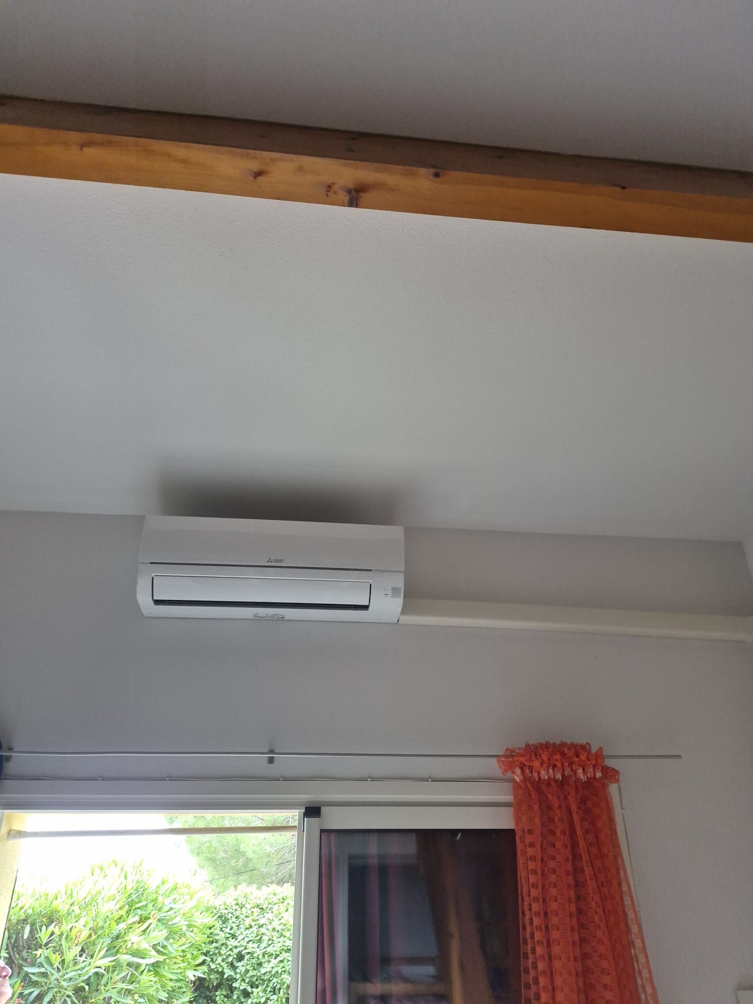 Climatiseur mural blanc au-dessus d'une fenêtre avec des rideaux orange et une poutre apparente au plafond.