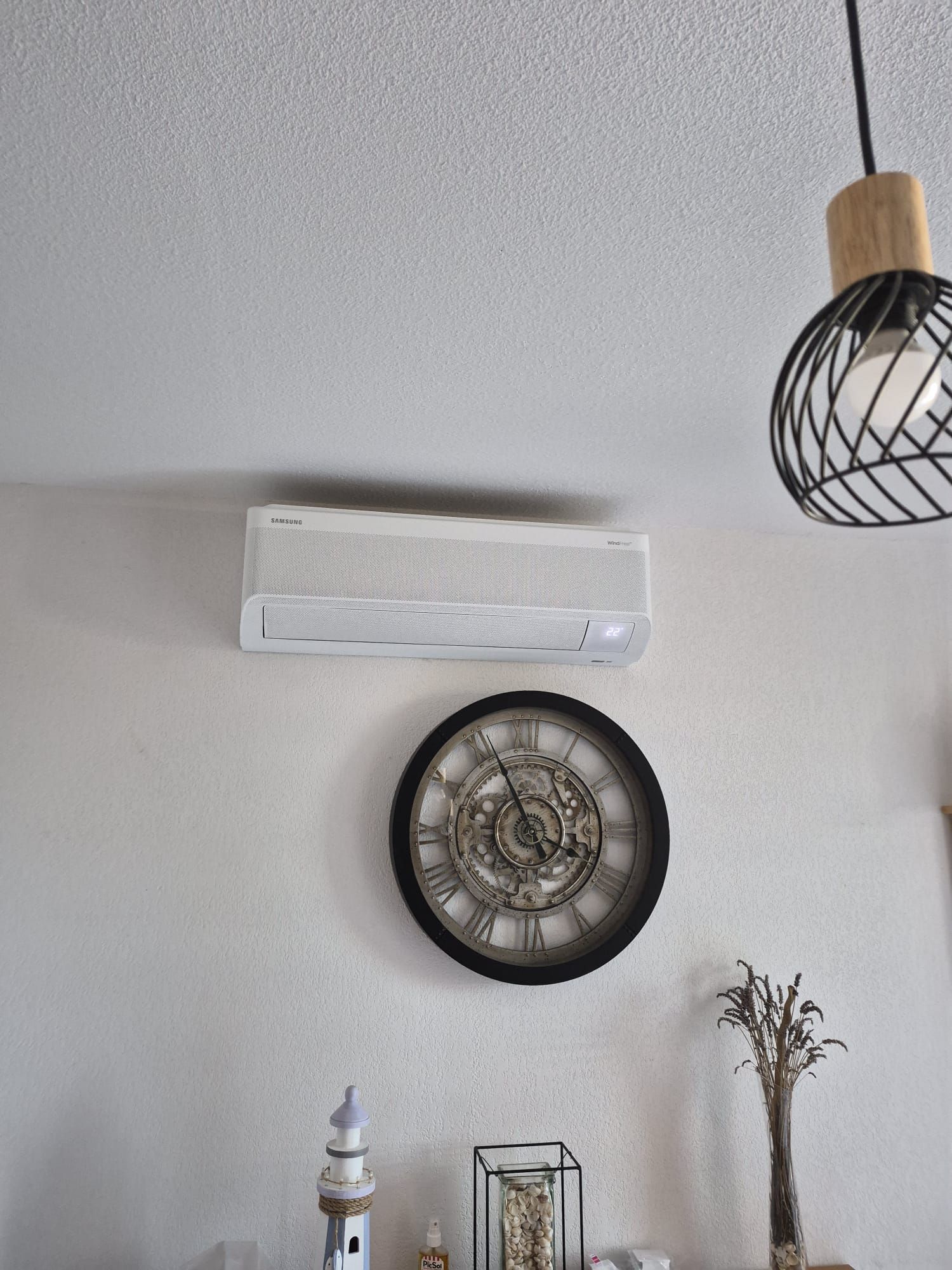 Mur blanc avec climatiseur mural, horloge murale ronde, lampe suspendue et petite étagère décorative.
