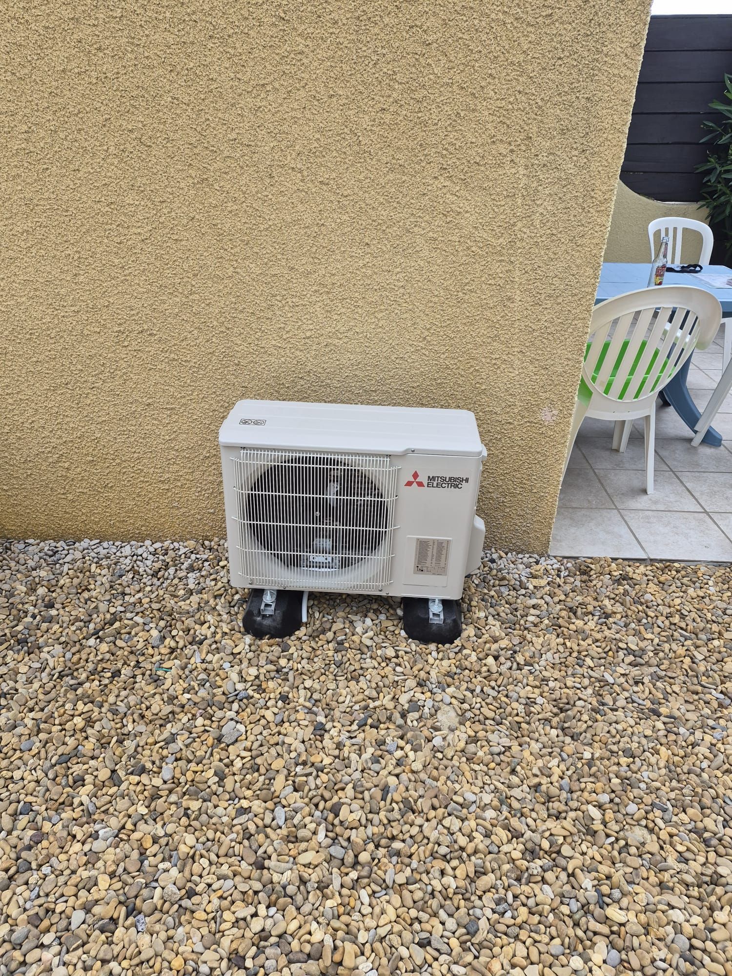 Unité de climatiseur extérieure contre un mur jaune sur une terrasse en gravier à côté de chaises en plastique blanc