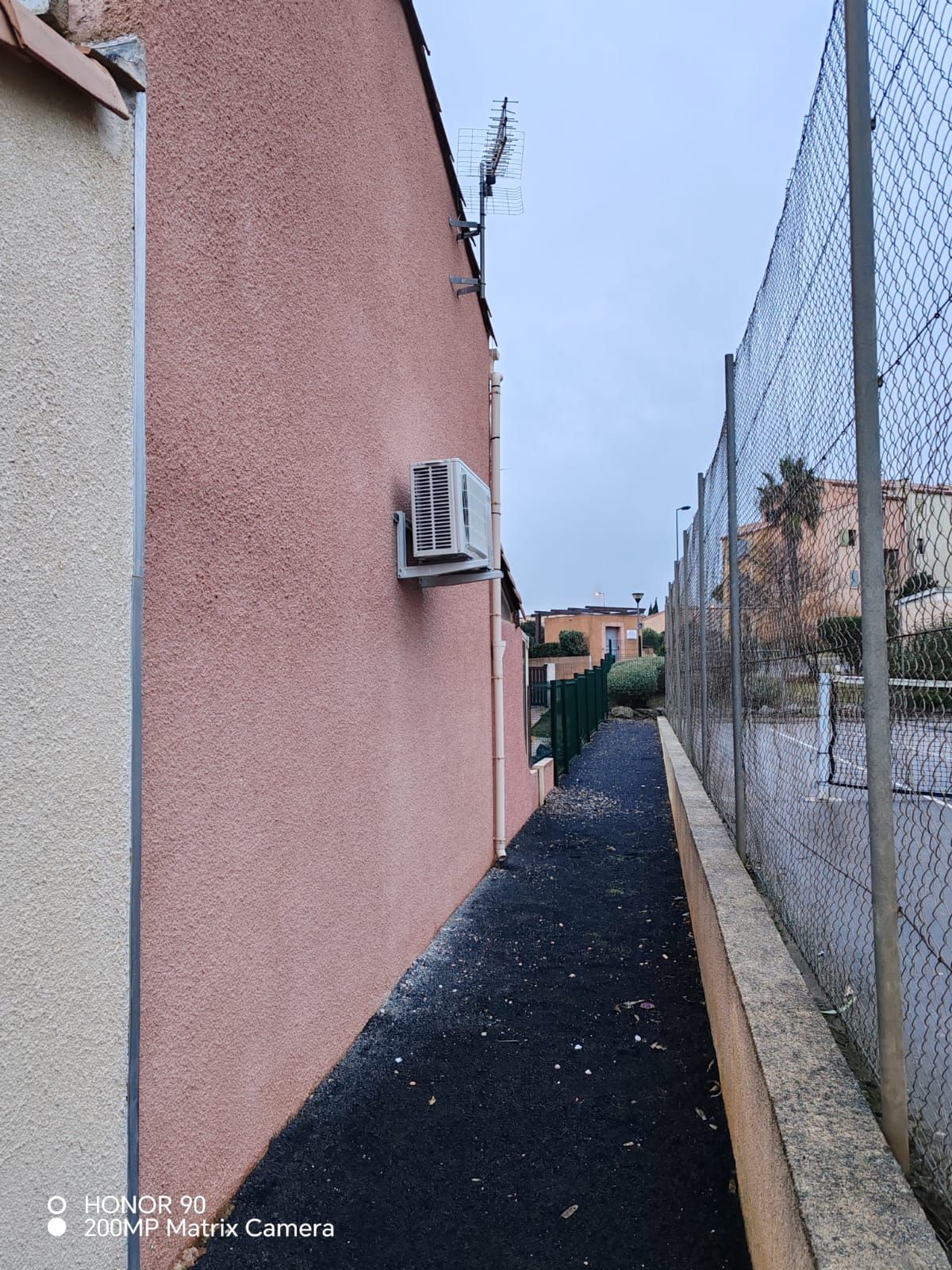 Ruelle étroite longeant un mur rose et une clôture en grillage, avec un climatiseur extérieur et des bâtiments au loin.