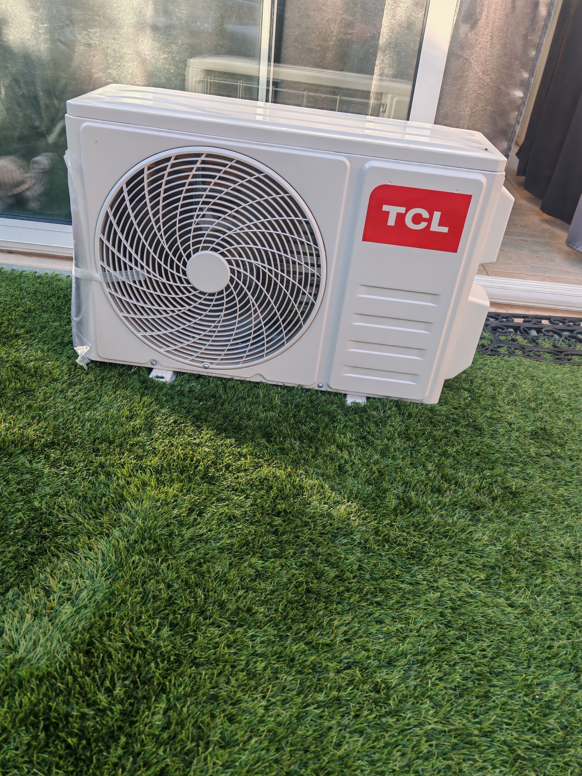 Unité de climatisation extérieure TCL posée sur l'herbe à côté d'une fenêtre.