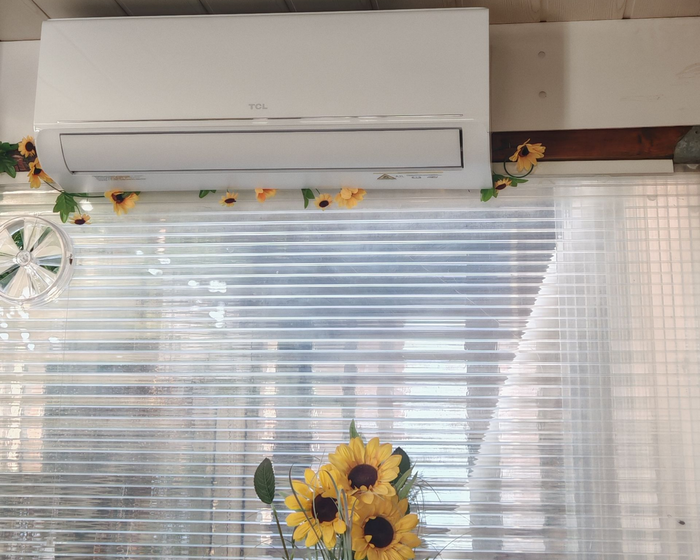 Climatiseur mural blanc au-dessus d'une fenêtre à stores, décorée d'une guirlande de tournesols et de tournesols en dessous.