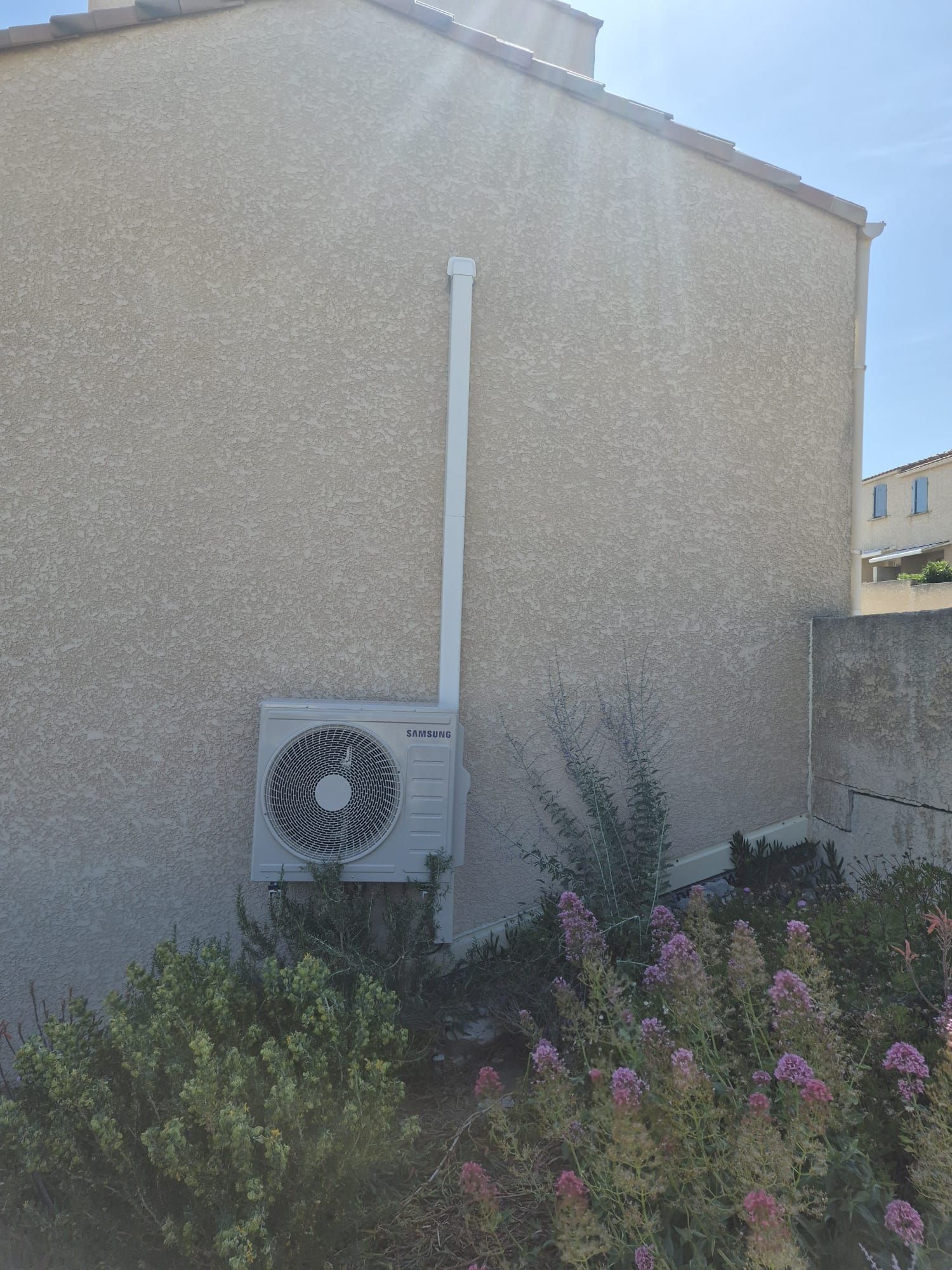 Extérieur d'une maison beige avec un climatiseur et des arbustes fleuris devant.