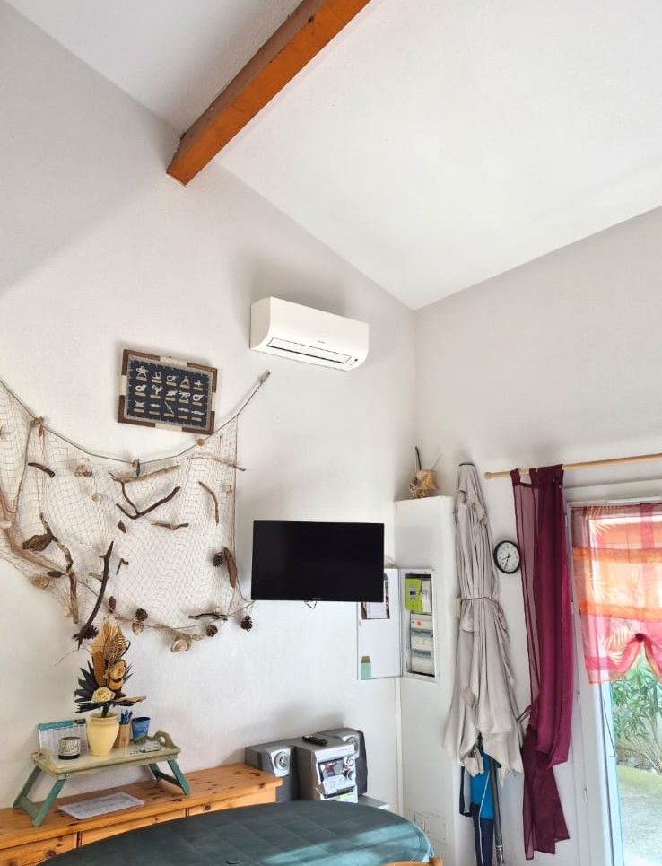 Petit coin salon avec télévision murale, climatiseur, étagère, rideaux et voilage décoratif.