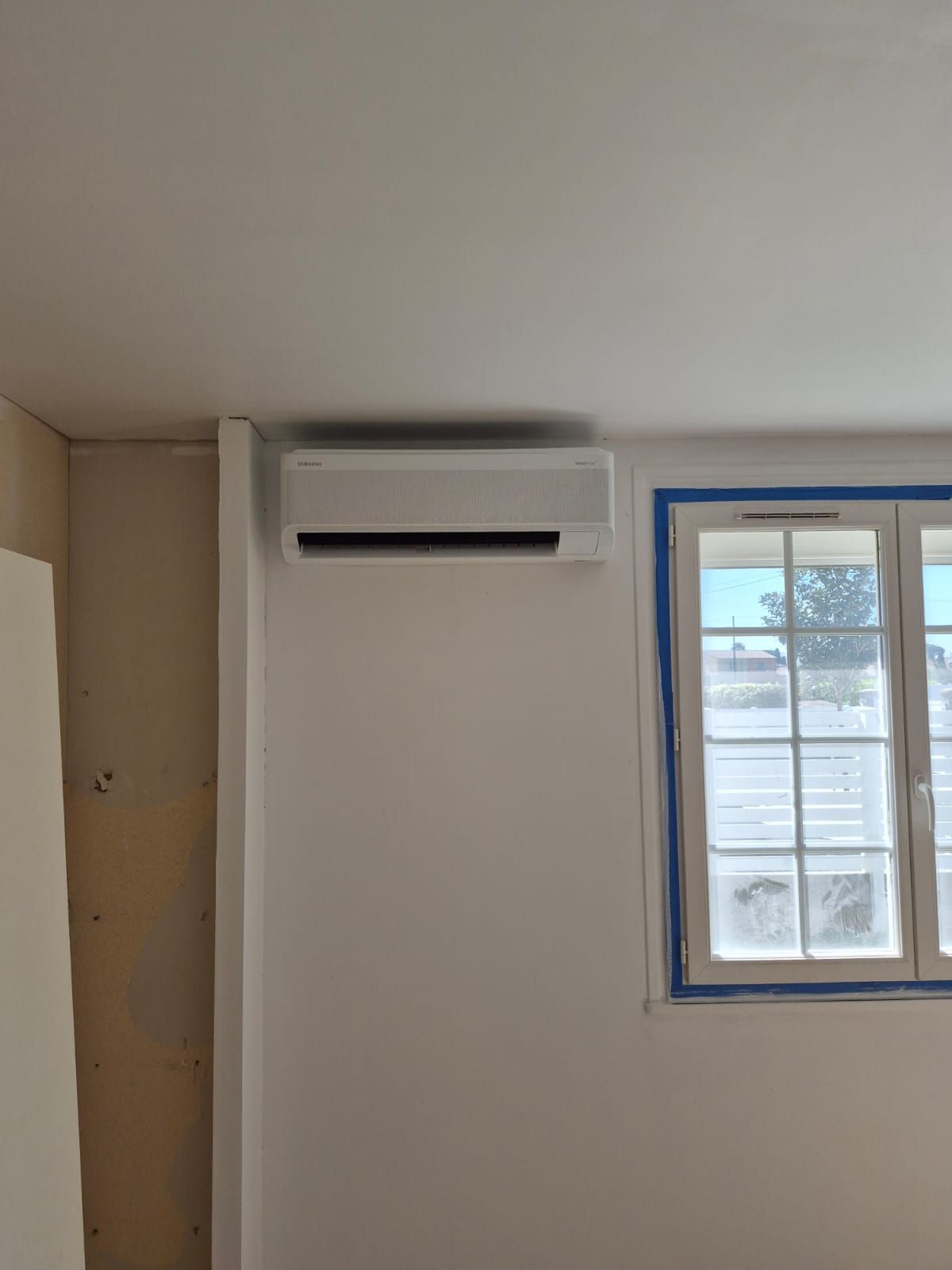 Climatiseur mural au-dessus d'une fenêtre à encadrement bleu dans une pièce sobre en cours de rénovation