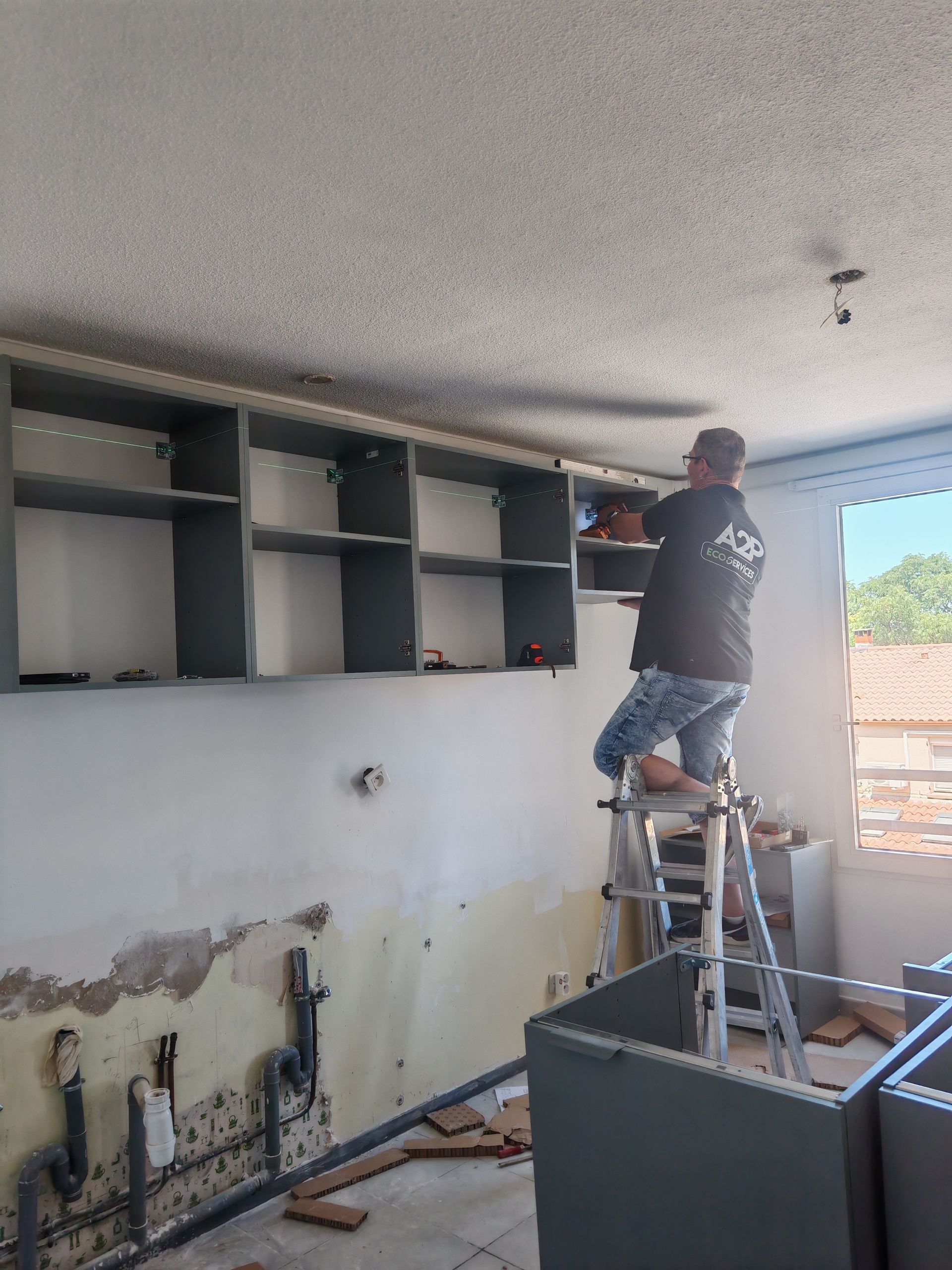 Une personne, debout sur une échelle, installe des armoires de cuisine hautes dans une pièce partiellement rénovée.