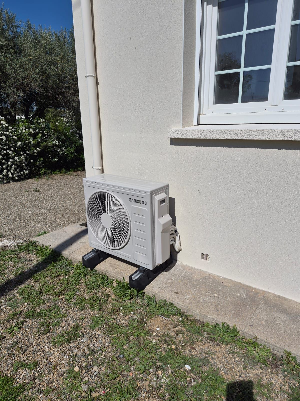Unité de pompe à chaleur extérieure située à côté d'un mur et d'une fenêtre d'une maison blanche, sur une bande de gazon.