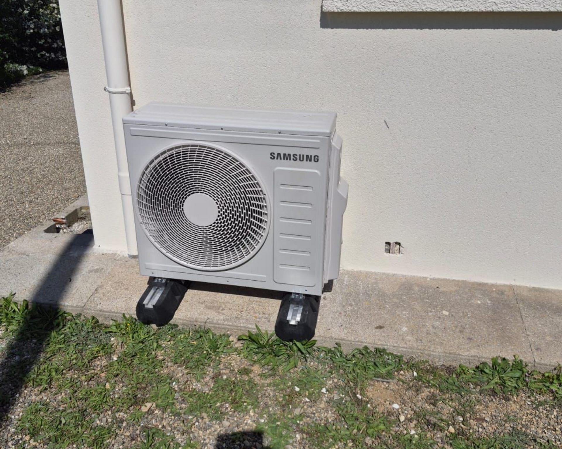 Unité de climatisation extérieure Samsung sur un support à côté d'un mur de maison et de l'herbe.