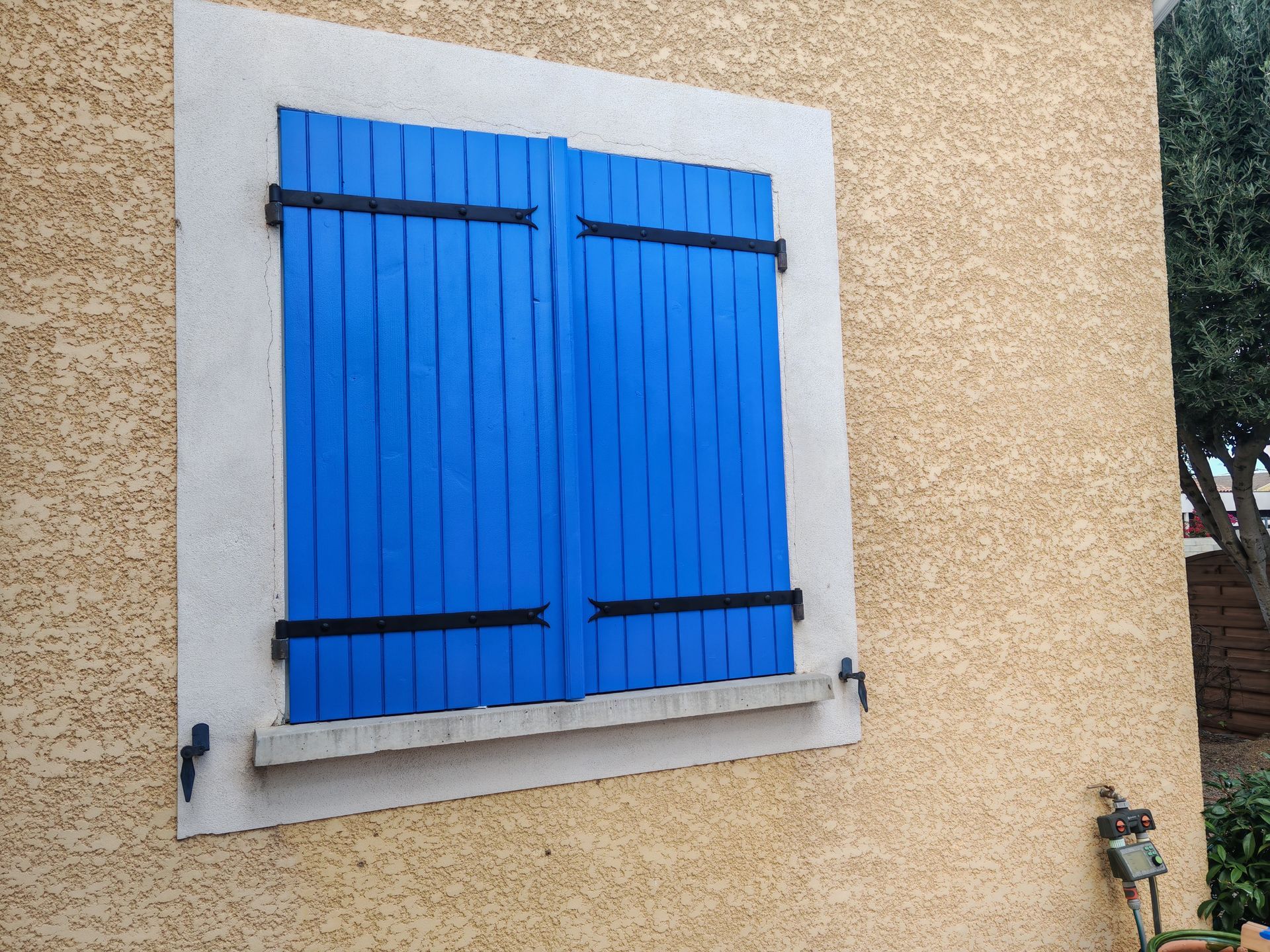 Volets bleus fermés sur un mur en stuc beige avec encadrement de fenêtre blanc
