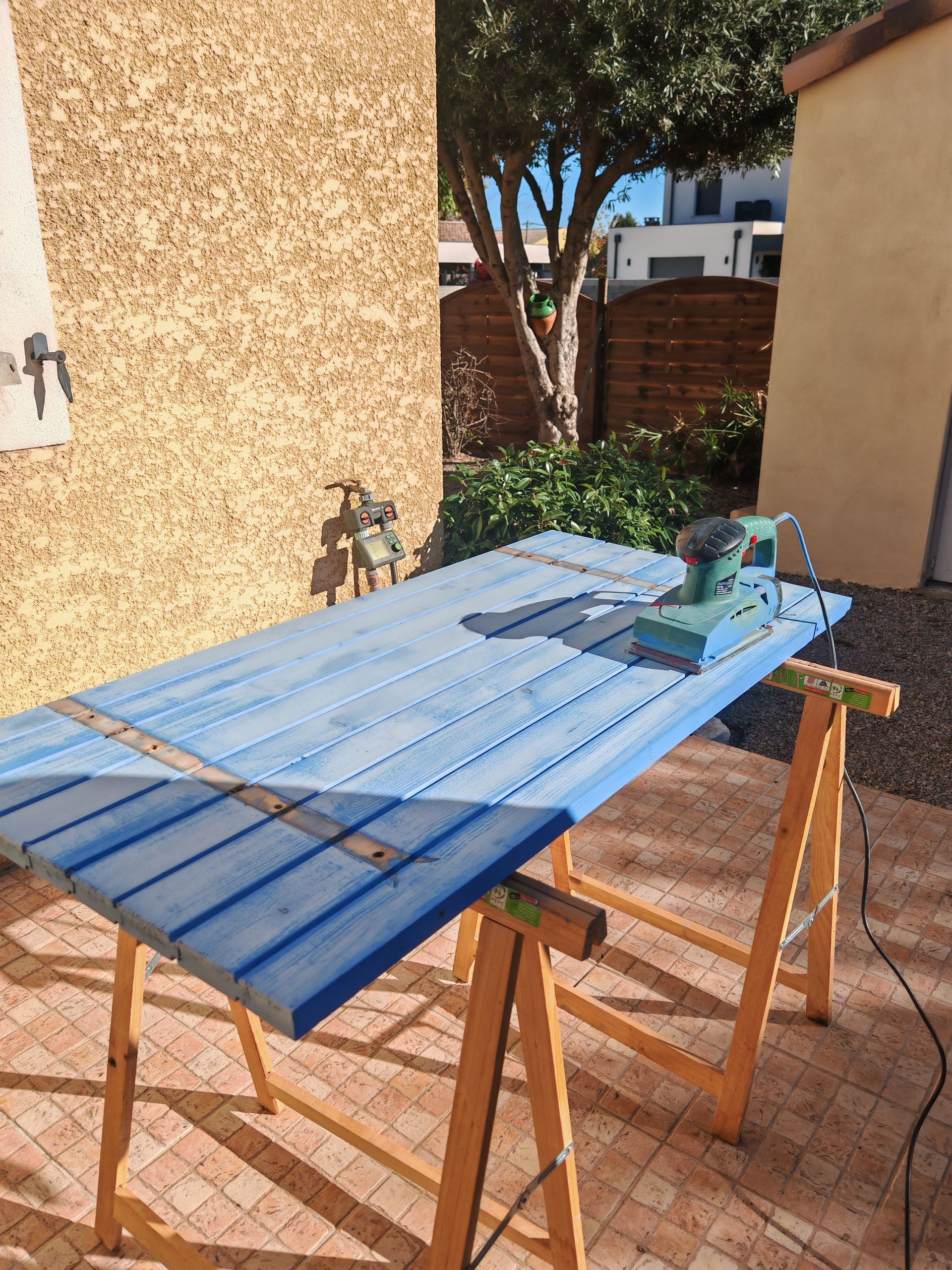 Des volets en bois bleus posés sur des tréteaux à l'extérieur, avec un outil électrique et des serre-joints.