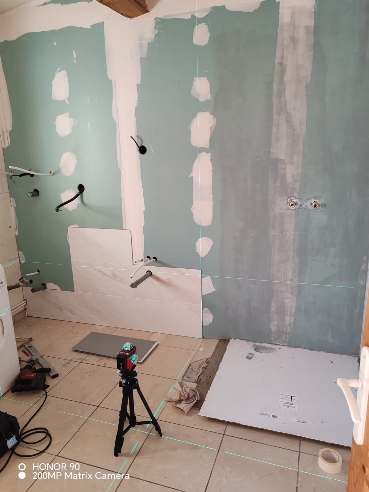 Murs de salle de bains en cours de rénovation avec des cloisons sèches, des câbles apparents, un sol carrelé et un trépied.