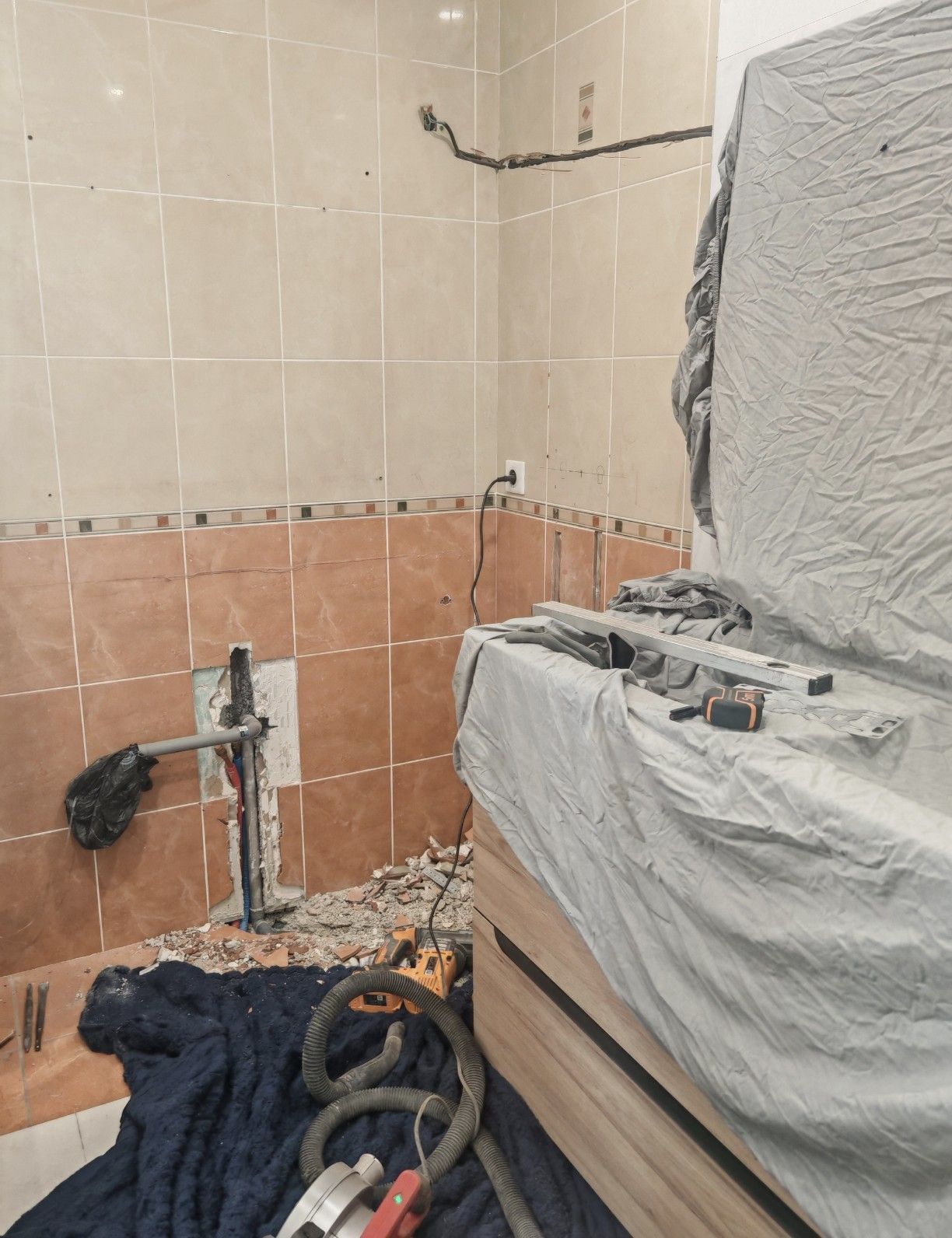 Salle de bains carrelée en cours de rénovation, avec plomberie apparente, outils et baignoire recouverte.